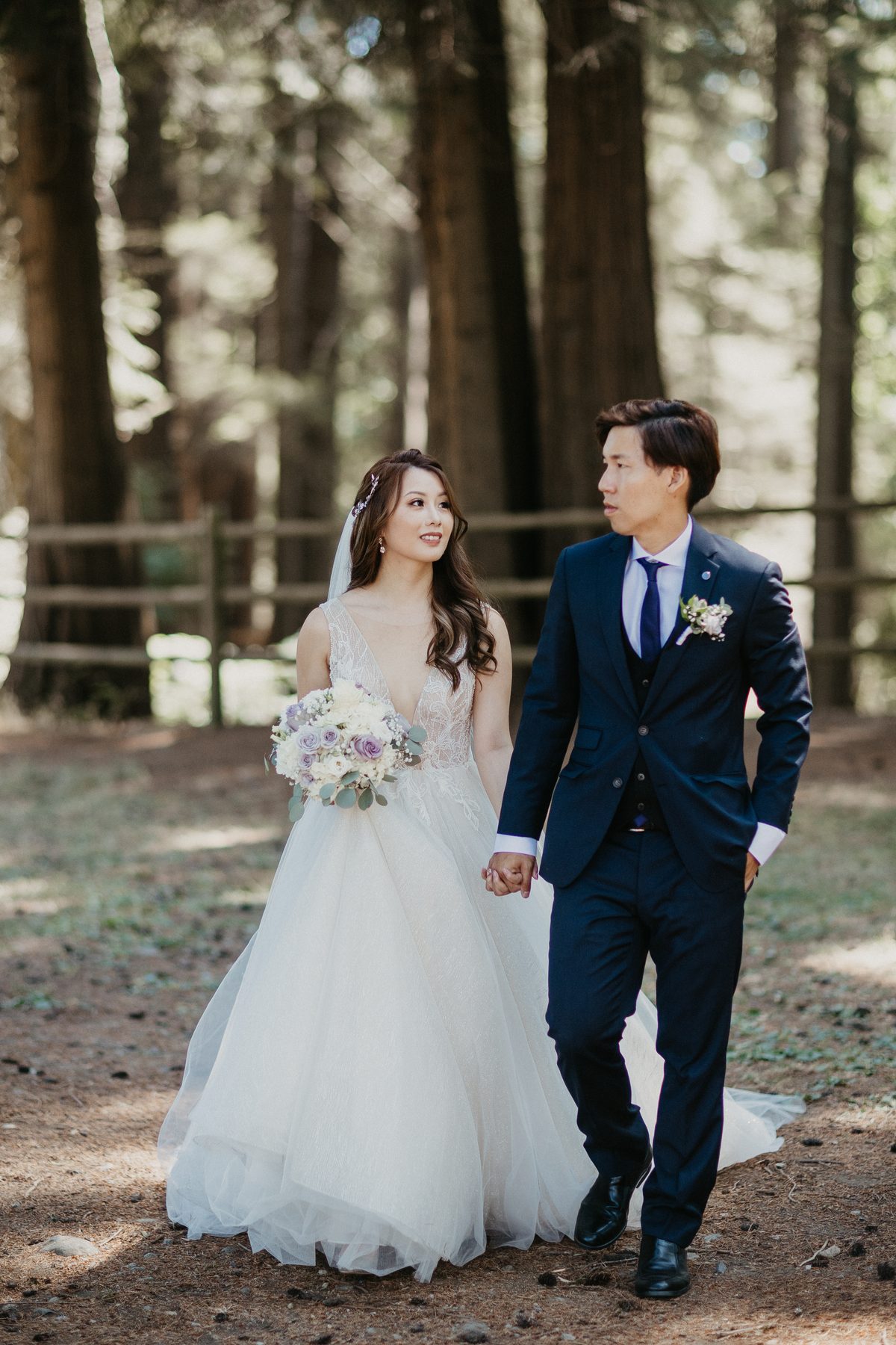 Couple walking hand in hand through the trees, the bride holding her bouquet