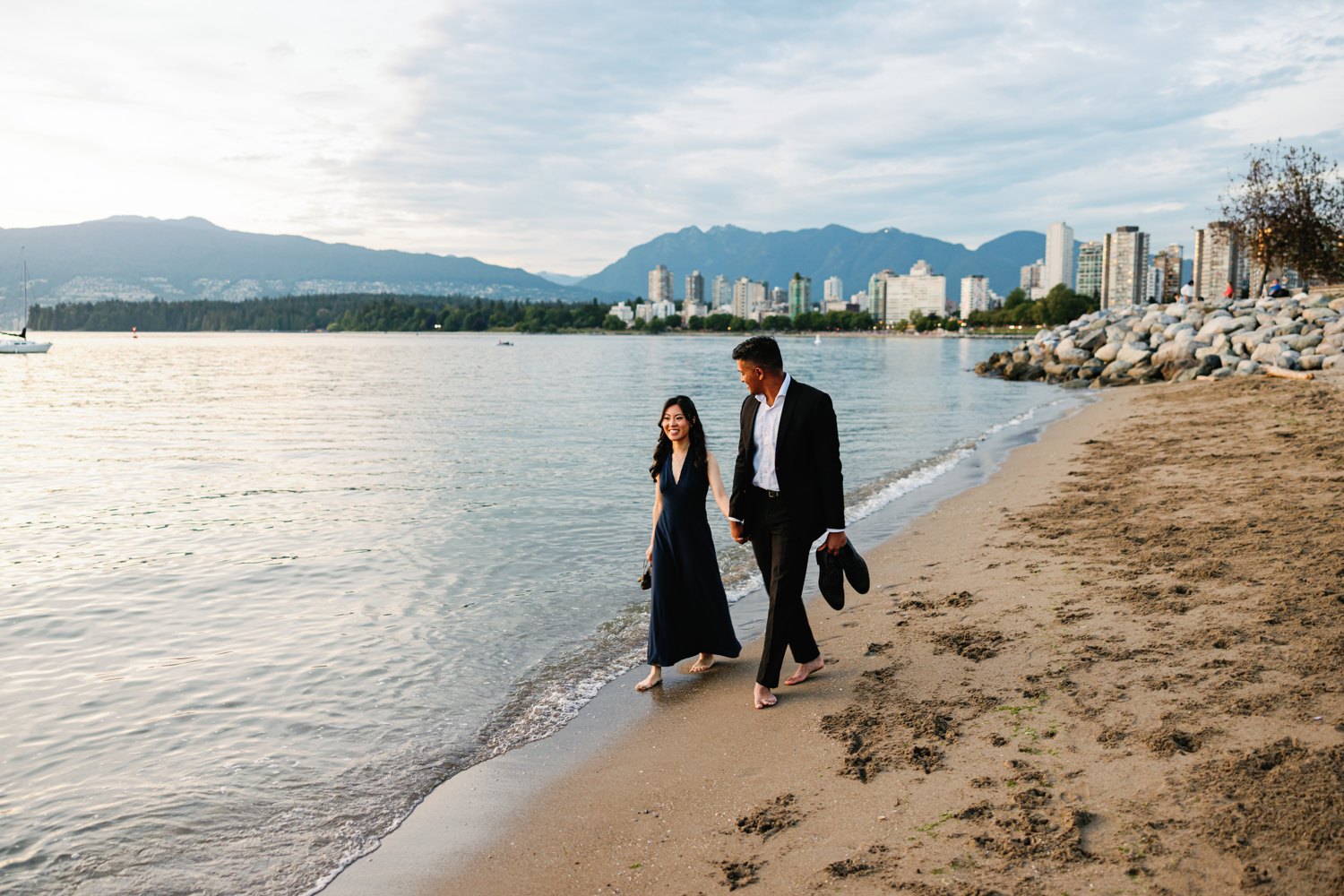 Eunice and Ashan walking a Kitsilano beach at golden hour, the city skyline across English Bay