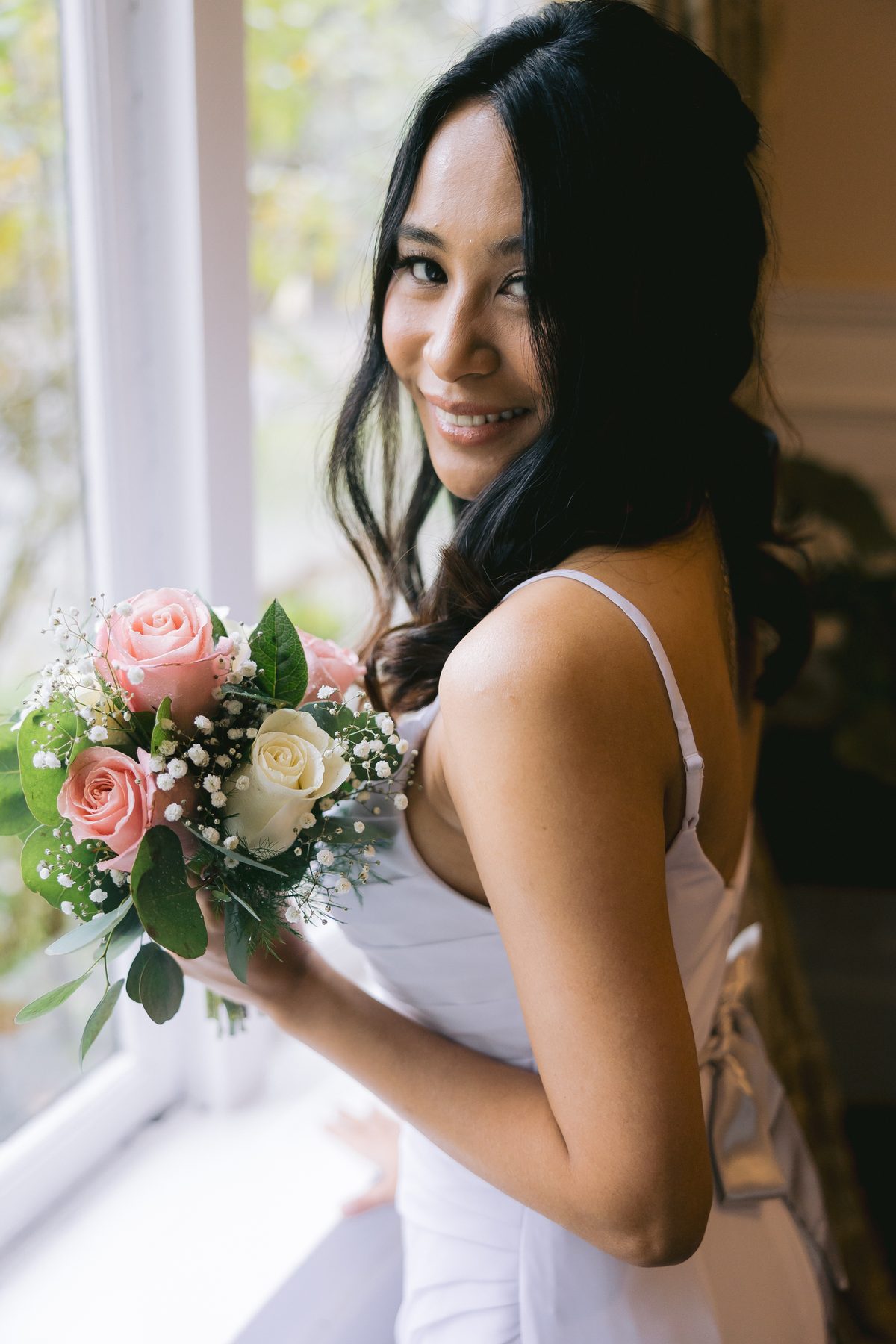 Bride in a white slip dress holding a pale rose bouquet, turned toward soft window light