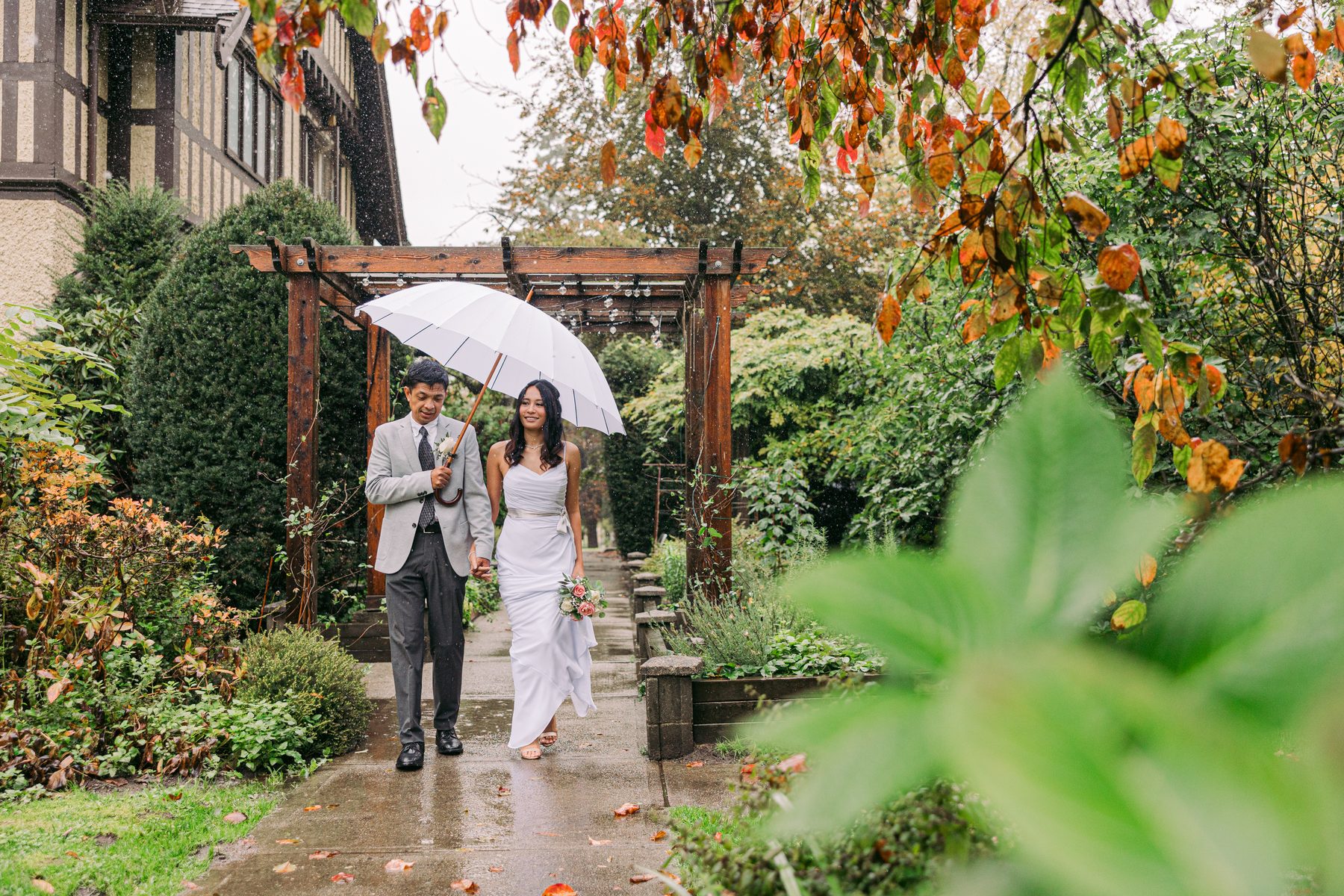 Bride and groom walking a wet garden path under a single white umbrella with autumn leaves overhead