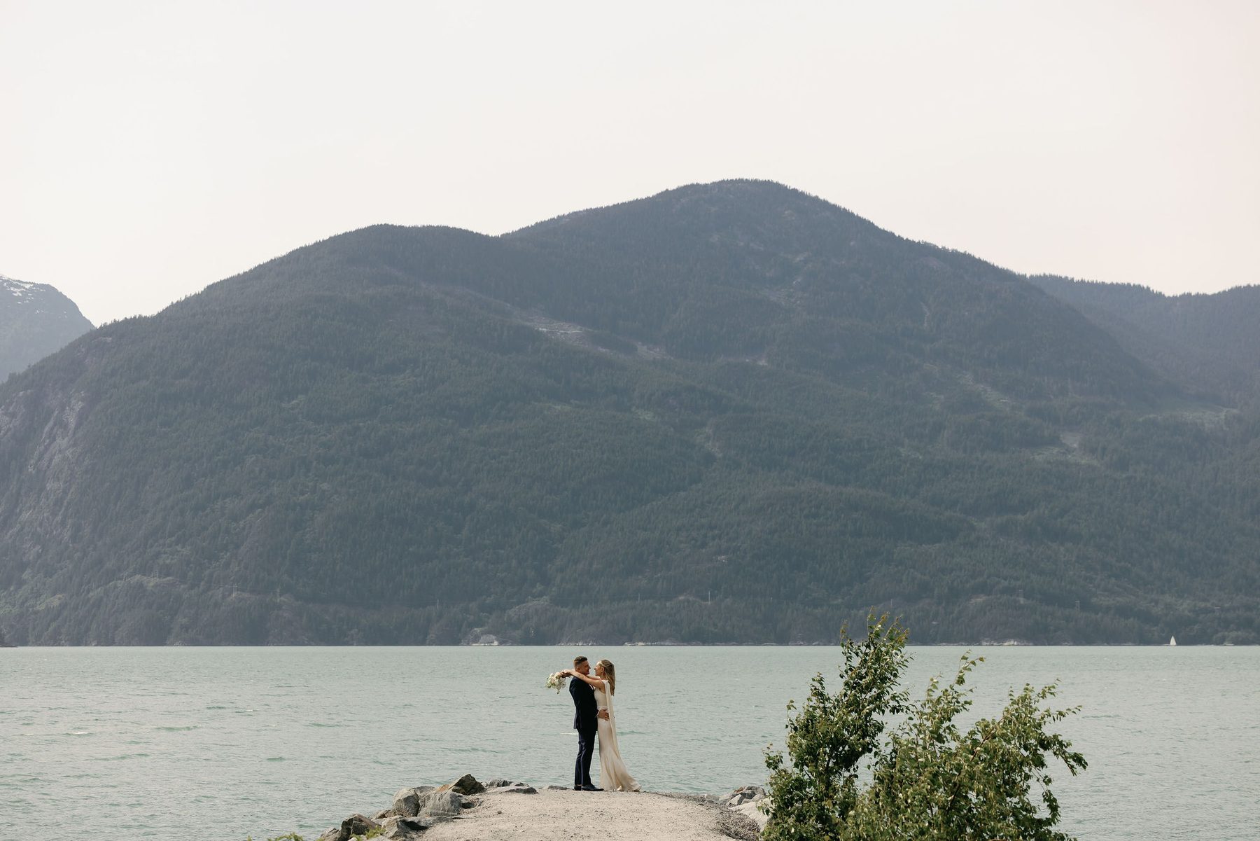 Tiny figures of a couple on a rock outcrop against a wide mountain and ocean backdrop