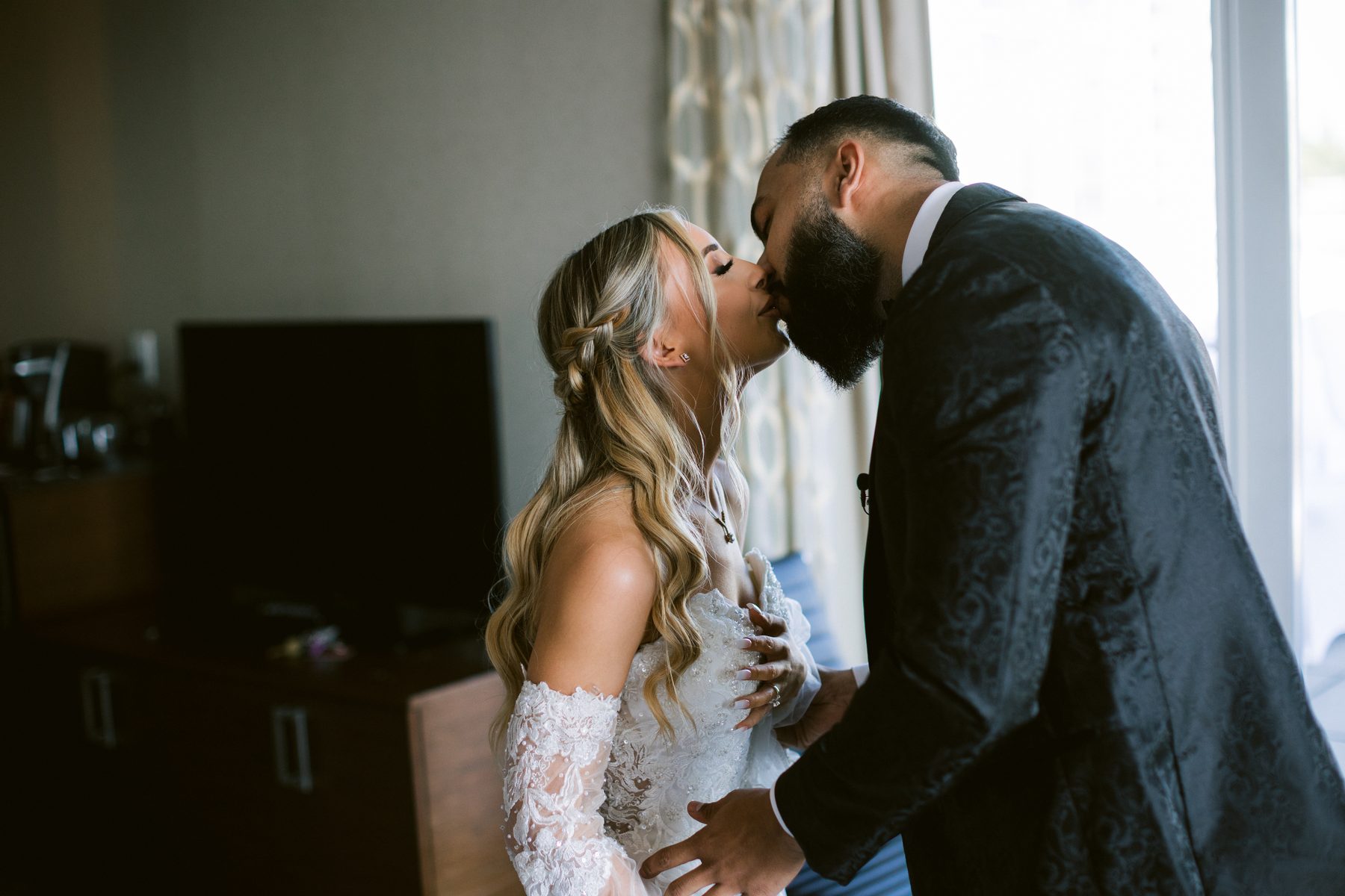 Groom leaning in to kiss the bride's forehead in a hotel suite with daylight coming through the window