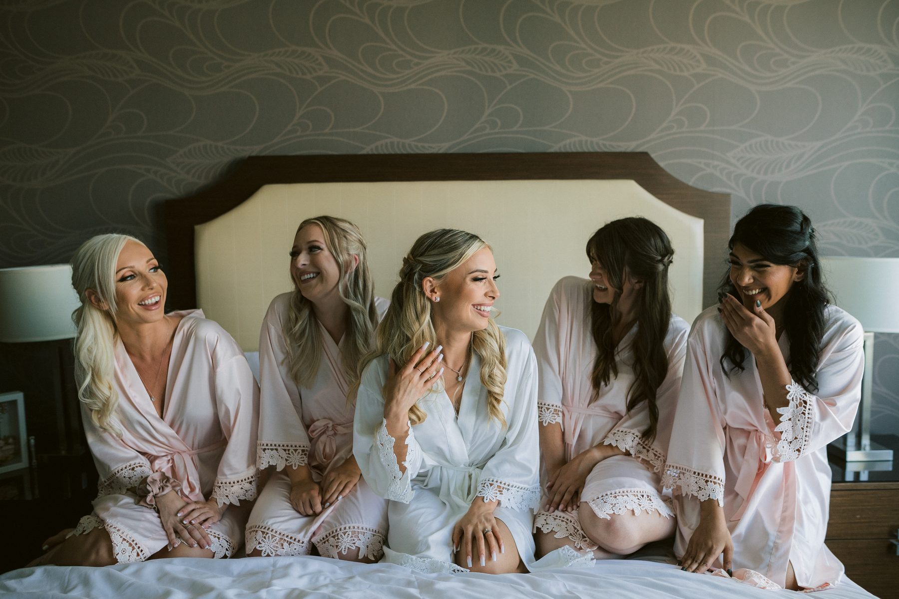 Five women in pale robes sitting together on a hotel bed, laughing, while the bride gets ready