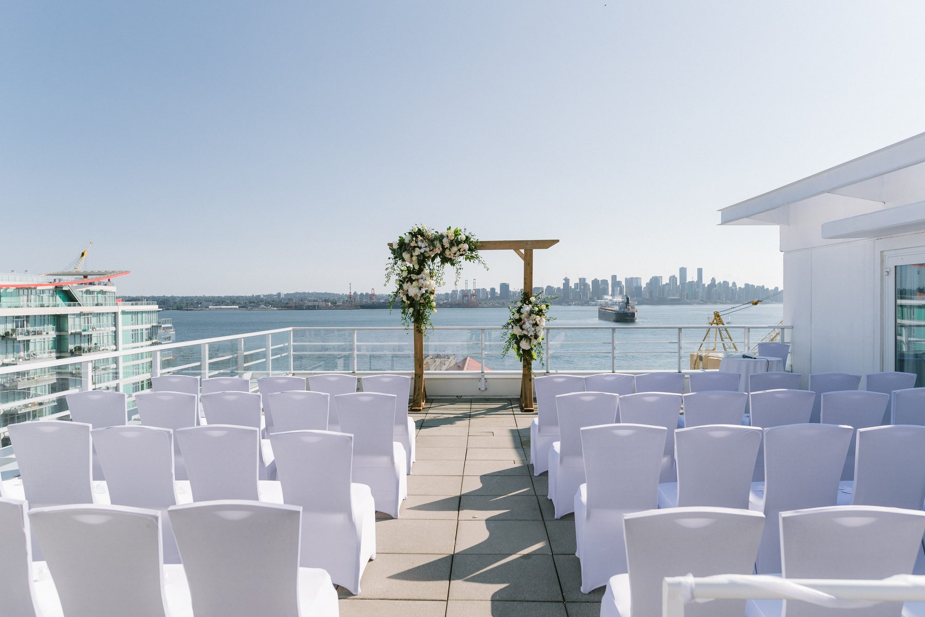 Rows of white ceremony chairs on a rooftop deck facing a timber arbour dressed in white and green florals