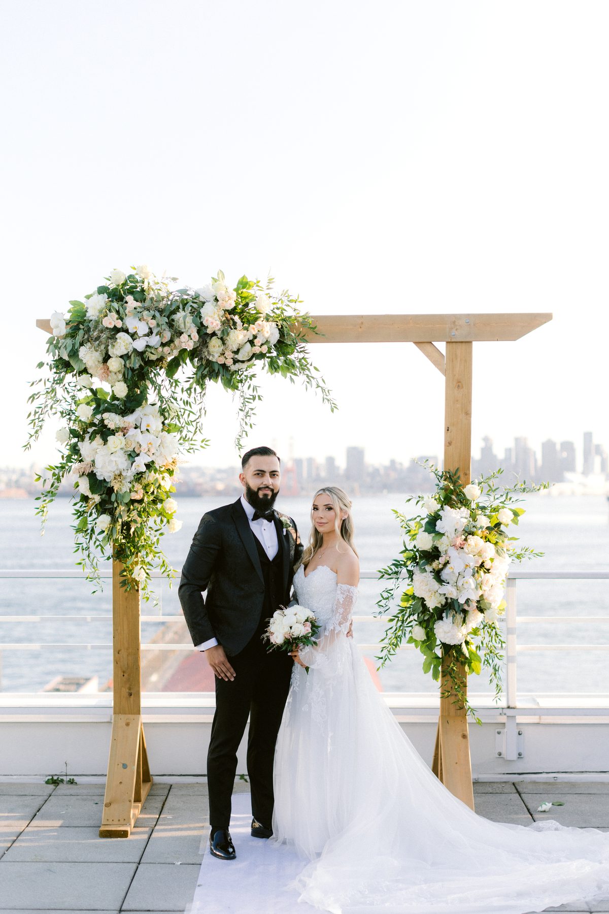 The couple standing under the floral arbour with the city skyline soft in the distance behind them