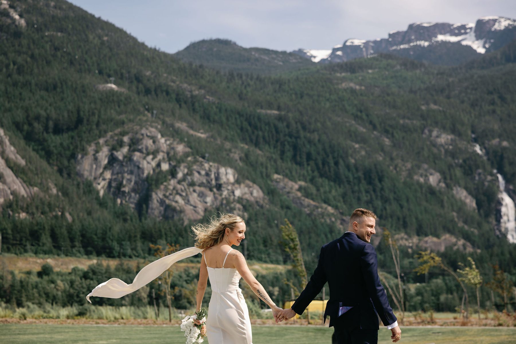 Bride's veil catching the wind as she and her groom walk with mountains behind them