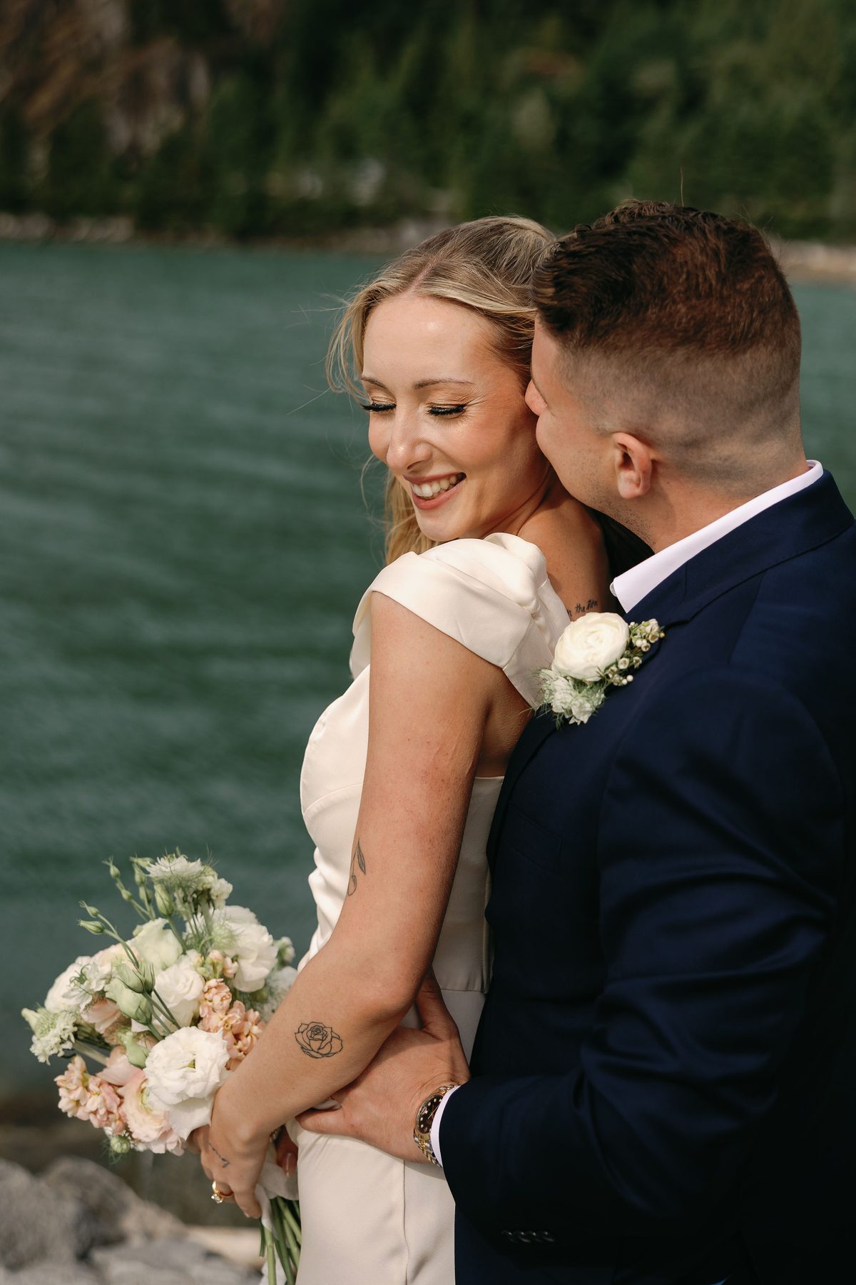 Bride laughing as her groom holds her from behind beside the water