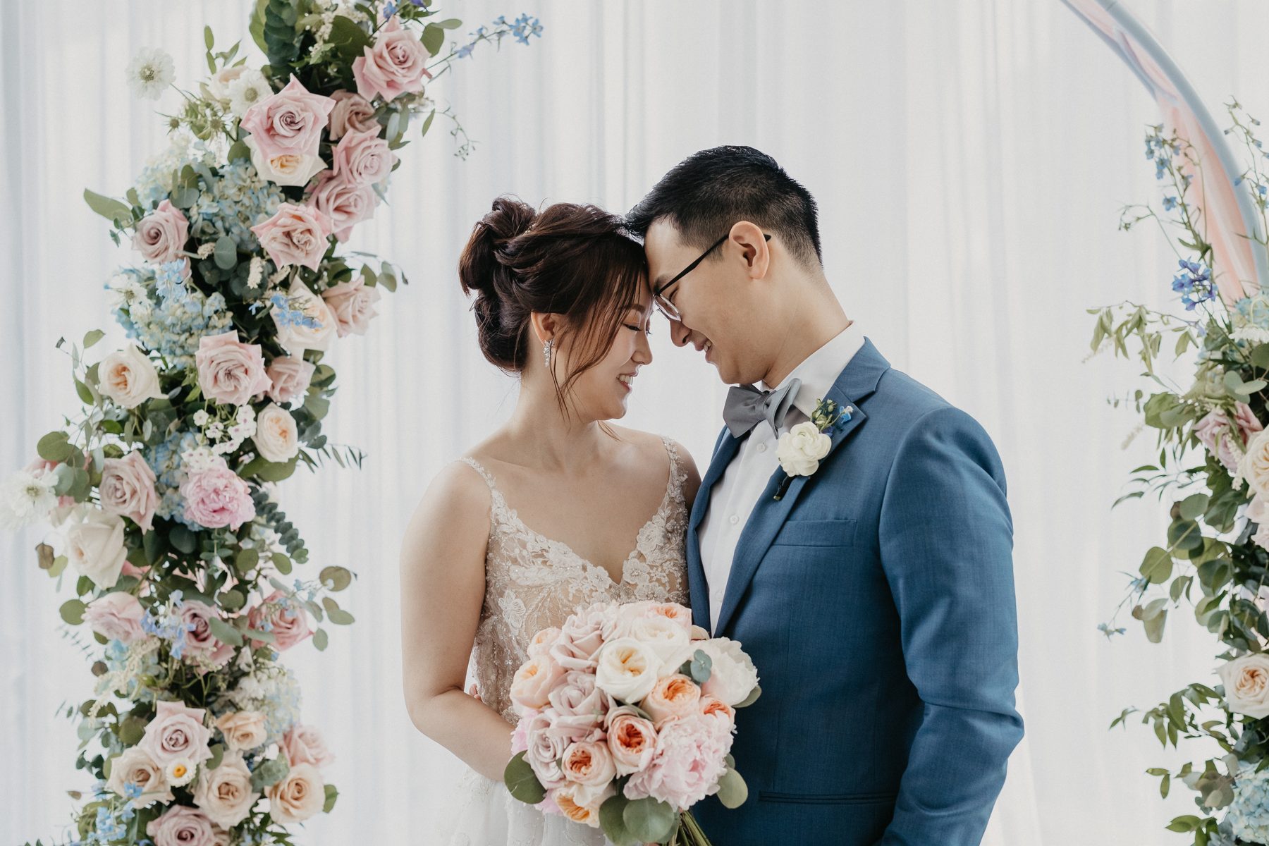 Chelsea and Andrew resting their foreheads together beside a floral arch at the Fairmont Pacific Rim