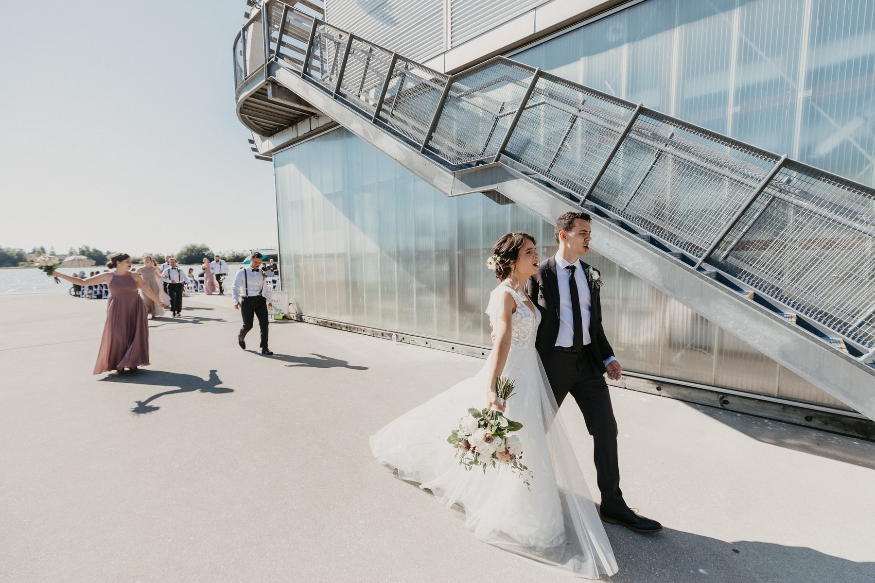 Bride and groom walking across a concrete dock past the glass wall of a riverside boathouse
