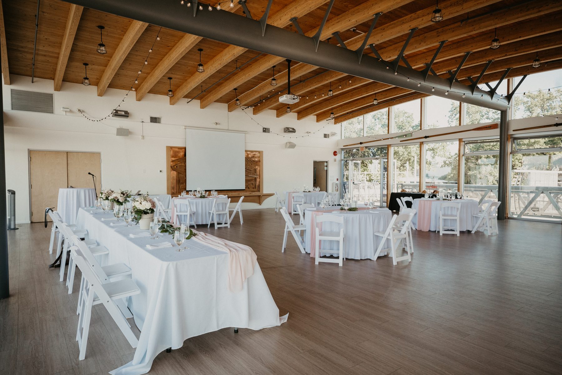 Reception hall with exposed timber beams and long glass windows overlooking the river, tables set in white